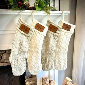 May include: Four cream-colored cable knit Christmas stockings hang from a white mantel. Each stocking has a brown leather patch with a name: Daddy, Mama, Austin, and Mallory. The stockings are decorated with a green and red garland.