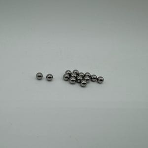 Pode incluir: Um grupo de pequenas bolas de metal prateadas, algumas estão dispostas em um aglomerado, enquanto duas estão separadas no lado esquerdo da imagem.