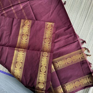 May include: A deep maroon fabric textile featuring a wide border with intricate gold-colored woven patterns of peacocks and circular motifs. The fabric includes thin gold-colored vertical stripes and finished edges with small tassels.