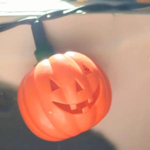 Puede incluir: Luz de calabaza de Halloween de plástico naranja con una cara sonriente. La luz tiene ojos triangulares, una sonrisa con dientes y una base verde. La luz está unida a un cable negro.