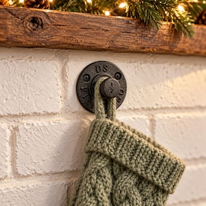 May include: A sage green cable-knit stocking hangs from a black metal pipe-style wall hook. The circular mounting plate features the letters DS and the number 1/2 embossed on the front, secured to a white painted brick wall surface.
