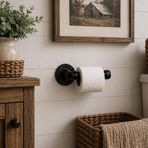 May include: A black industrial-style toilet paper holder mounted on a white wall. The holder has a round base and a cylindrical arm holding a roll of white toilet paper. A framed picture and a woven basket are also visible.