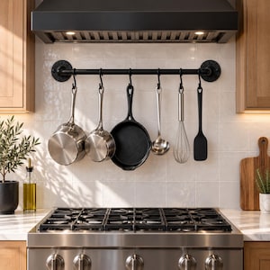 May include: A black industrial-style pipe wall rack mounted above a stove, featuring six S-hooks holding two stainless steel saucepans, a black cast iron skillet, a stainless steel ladle, a wire whisk, and a black silicone spatula.