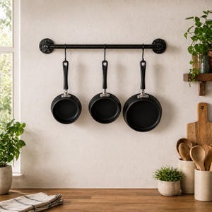 May include: Three black non-stick frying pans hanging from a black metal bar on a kitchen wall. The pans have black handles and are suspended by hooks. The kitchen setting includes wooden countertops, plants, and a window.