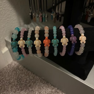 May include: A set of ten colorful beaded bracelets with a turtle charm on each. The bracelets are displayed on a black surface.