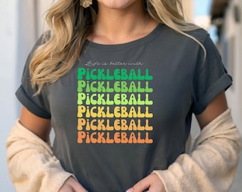 May include: A dark grey t-shirt with the text "Life is better with" and the word "Pickleball" repeated five times in a rainbow gradient. The text is in a retro font.