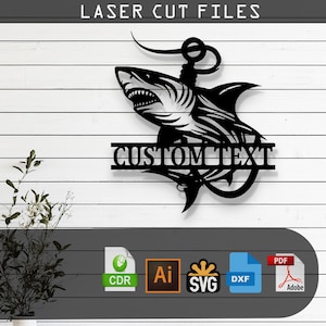 May include: Black metal laser-cut wall art featuring a shark design with an anchor and the words "CUSTOM TEXT". The artwork is mounted on a white wooden plank background. The top of the image reads "LASER CUT FILES".
