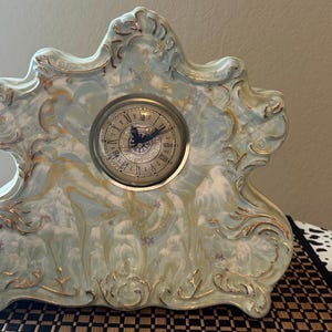 May include: An antique-style mantel clock with a light blue and gold marbled design. The clock face is round with Roman numerals and ornate hands. The clock's decorative frame has a scrolled design.