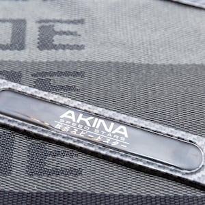 Può includere: Una piastra in metallo nero e argento con il testo "AKINA SPEED STARS" e caratteri giapponesi. La piastra ha un motivo in fibra di carbonio.