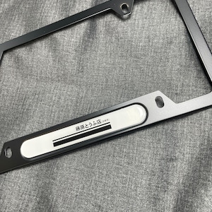 May include: A dark grey license plate frame with a rectangular white section. The white section has black Japanese text. The frame has mounting holes and a sleek, modern design.