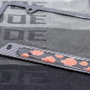 May include: A black carbon fibre license plate frame with a red and black graphic of stylised clouds. The frame has a textured surface.