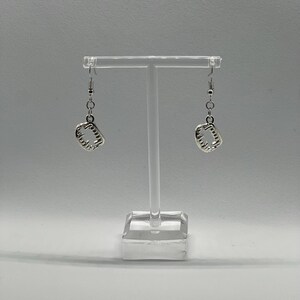 May include: A pair of silver dangle earrings with a textured square design. The earrings are hanging from a clear display stand.