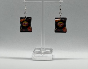 May include: A pair of dangle earrings featuring a miniature Lay's potato chip bag design. The earrings are silver and the bag is black with red and yellow accents. The bag has the word "Lay's" printed on it.
