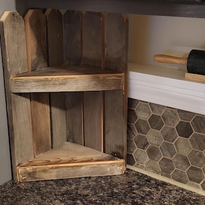 Puede incluir: Una estantería de esquina de madera rústica con dos estantes. La estantería está hecha de madera recuperada y tiene un acabado natural y desgastado. La estantería es perfecta para exhibir objetos pequeños o plantas.