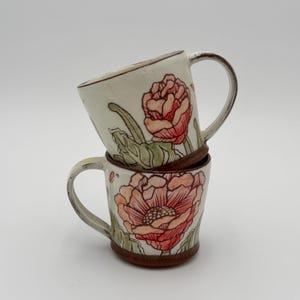 May include: Two ceramic mugs stacked on top of each other. Each mug features a hand-painted floral design with pink and red flowers, green leaves, and brown accents. The mugs have a white background and a curved handle.