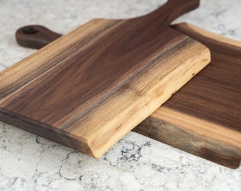 May include: Two rectangular wooden serving boards made from dark walnut wood with natural live edges. Each board features a smooth, polished surface showing the wood grain and a handle with a circular hole for hanging, arranged in an overlapping position.