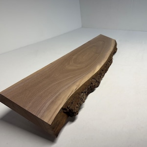 May include: A rectangular, unfinished wooden slab with a natural edge. The wood has a rich brown color with visible grain patterns. The live edge is dark brown and textured.
