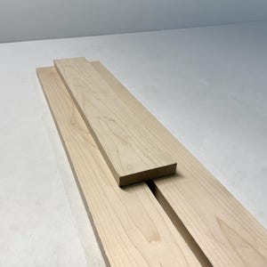 May include: Three unfinished wooden boards, likely for woodworking projects. The boards are a light, natural wood color with visible grain patterns. One board is stacked on top of the other two.