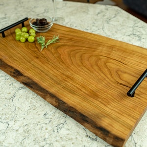 May include: A large wooden serving tray with live edge details and two black metal handles. The tray is made of light brown wood with a natural finish. There are green grapes and a small bowl of olives on the tray.