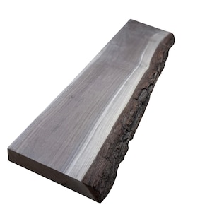 May include: A rectangular, gray wooden slab with a natural, live edge. The wood grain is visible, and the bark edge is dark brown. The slab is likely for woodworking or crafting projects.