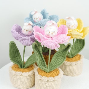 May include: Four crocheted flower pots with bunny faces. Each pot is a different colour: pink, purple, blue, and yellow. The pots are sitting on a white surface.