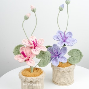 May include: Two crocheted flower arrangements in pots. One arrangement has pink flowers and the other has purple flowers. Both arrangements have green leaves and are in beige pots with brown rims.