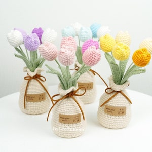May include: A bouquet of crocheted tulips in various pastel colours, including pink, purple, yellow, and blue, arranged in small crocheted bags with brown ribbon ties. Each bag has a brown label with the text "Keep your smile".