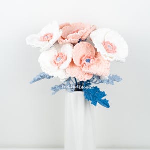 May include: A bouquet of crocheted flowers in white and pink with blue crocheted leaves in a white vase. The flowers are arranged in a vase and the text "Knot Knot Bloom" is visible in the image.