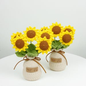 May include: Two crochet sunflower bouquets in cream-coloured vases. Each bouquet features yellow petals, brown centres, and green leaves. The vases have a brown string tie and a tag that reads "keep your smile".