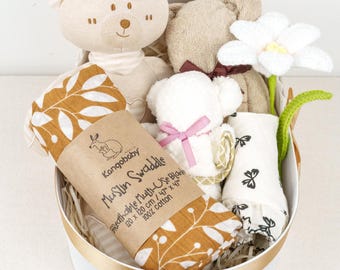 Può includere: Un cesto regalo bianco pieno di articoli per bambini, tra cui una copertina in mussola marrone e bianca con un motivo a foglie, un orsacchiotto bianco, un orsacchiotto marrone, un fiore bianco e un panno per bambini bianco e grigio. Il cesto è decorato con un nastro dorato.