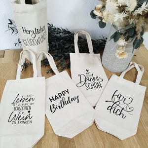 Könnte beinhalten: Eine Auswahl an cremefarbenen Canvas-Tragetaschen mit schwarzer Schrift. Eine Tasche trägt die Aufschrift "Happy Birthday", eine andere "Danke", und weitere haben deutsche Sätze. Eine Weinflasche befindet sich in einer Tasche. Trockenblumen in einer Vase.