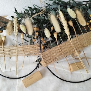 Peut inclure: Deux couronnes circulaires en métal noir décorées de blé séché, d'herbes beiges duveteuses et de capsules de graines. Le centre de chaque couronne est tissé avec de la ficelle naturelle. Un bouchon et de la verdure complètent le design rustique.