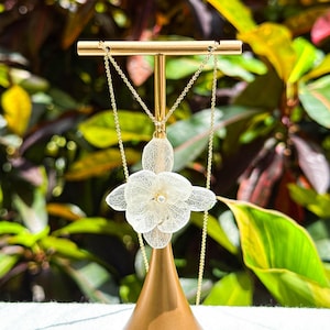 Puede incluir: Un collar dorado con un colgante blanco en forma de flor. El colgante presenta un diseño calado detallado y una pequeña perla. El collar se exhibe sobre un soporte dorado, sobre un fondo de hojas verdes y rojas.