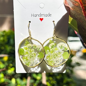 May include: Handmade gold-tone earrings with clear resin, featuring pressed white flowers and green leaves. The earrings have a circular design and are suspended from gold-coloured ear wires. The card behind the earrings says "Handmade" with a red heart.