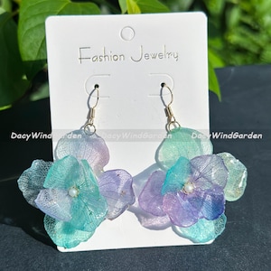 Op de afbeelding: Hangende oorbellen met gelaagde hortensia bloem ontwerpen in tinten van turquoise, paars en groen. De oorbellen hebben goudkleurige haken en een kleine parel in het midden van elke bloem. Ze worden gepresenteerd op een witte kaart met "Fashion Jewelry".
