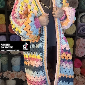 May include: A long, open-front crochet cardigan in a patchwork design. The cardigan features a vibrant mix of colours including pink, yellow, blue, orange, and grey. The sleeves are long and the garment is knee-length. Text on the image reads "AS SEEN ON TIKTOK @SELFMADECROCHET".