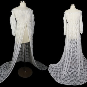 May include: A white lace off-the-shoulder gown with long sleeves and a flowing train. The dress is displayed on a mannequin against a black background, showcasing its delicate floral lace pattern and elegant design. The gown's open front adds to its ethereal look.
