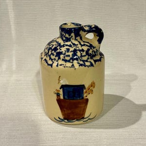 May include: A small, cream-colored ceramic pitcher with a blue and white crackle glaze. The pitcher has a brown and blue painted design of a house on a boat.