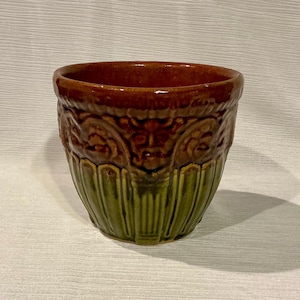 May include: A brown and green ceramic planter with a decorative design. The planter has a wide rim and a rounded base.