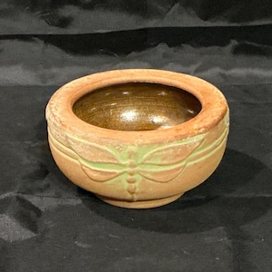 May include: A small, round, terracotta-colored bowl with a light green dragonfly design around the exterior. The interior of the bowl is a dark brown color. The bowl is likely made of ceramic or clay.