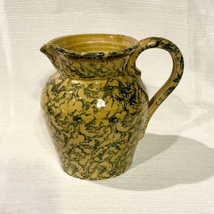 May include: A yellow ceramic pitcher with a green marbled design. The pitcher has a wide mouth and a handle.