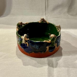 May include: A ceramic bowl with a dark green interior, encircled by small monkey figurines. The exterior features a band of blue and green, with a red base. The monkeys appear to be holding onto the rim of the bowl.
