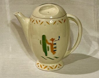 May include: A cream-colored ceramic teapot with a stylized cactus and cowboy design. The teapot has a lid with a small hole in the top and a handle on the side.