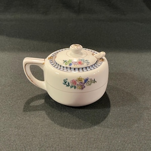 May include: A small, white ceramic sugar bowl with a lid and handle. The bowl features a floral design with blue, pink, yellow, and green accents. The lid has a matching floral pattern and a small knob. A small spoon rests on the lid.