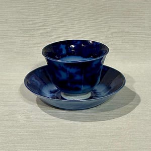 May include: A blue ceramic teacup and saucer set. The teacup has a faceted design and sits on a matching saucer. The set is a deep shade of blue, with a glossy finish. The teacup is approximately 3 inches in diameter.