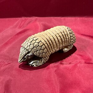 May include: A detailed figurine of an armadillo, featuring a textured, segmented shell in shades of tan and brown. The armadillo's head and feet are a metallic silver color, with visible claws. The figurine is set against a deep red background.