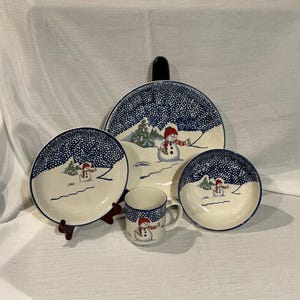 May include: A ceramic dinnerware set featuring a snowman motif with a red hat and scarf against a dark blue snowy background. The set includes a large dinner plate, a smaller plate, a bowl, and a mug, all decorated with matching winter scenes.