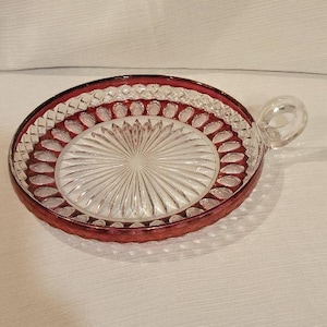 May include: A round clear glass coaster with a deep red rim and a small side handle. The center features a sunburst pattern of radiating lines, surrounded by a border of embossed teardrop shapes and a diamond-cut edge along the outer perimeter.