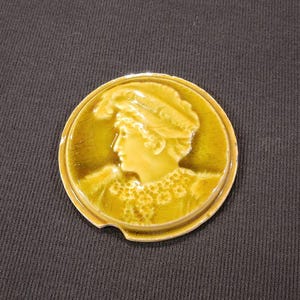 May include: A ceramic tile with a raised image of a woman wearing a hat and a necklace. The tile is a light brown color with a green glaze.