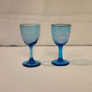 May include: Two matching blue stemmed glasses with etched designs. The glasses have a clear bowl and a dark blue stem and base. The etched design appears to be a landscape. The glasses are on a white surface.